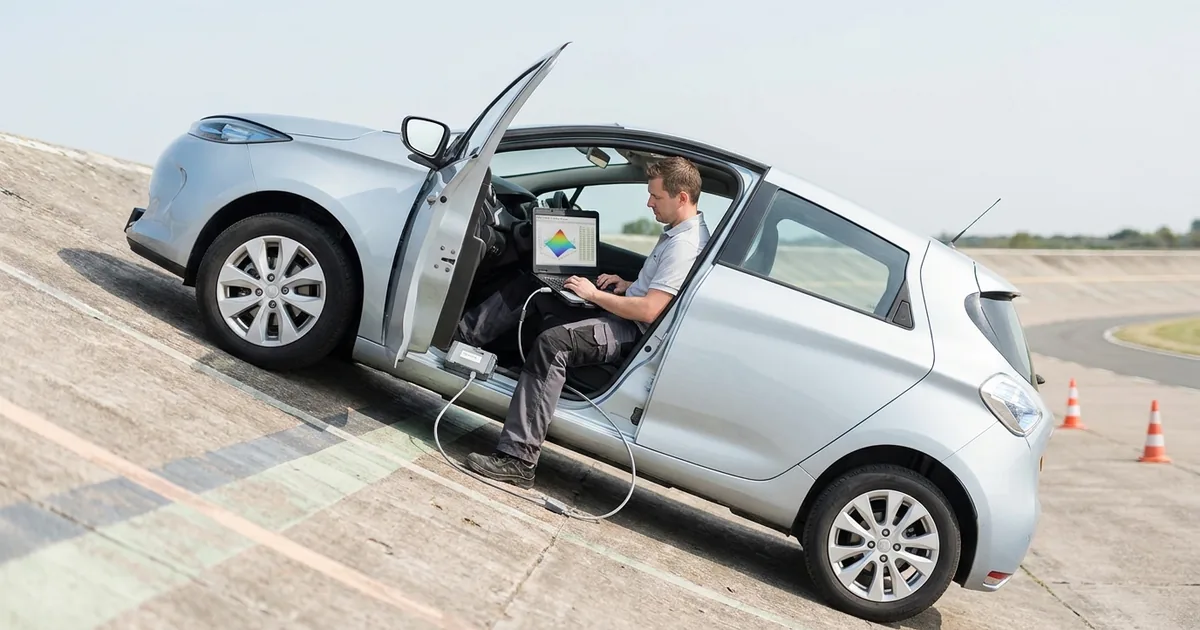 A calibration engineer sits in the open driver door of a silver hatchback parked on a proving-ground slope, tuning values on a laptop that shows a calibration map, cabled to an interface box on the door sill.