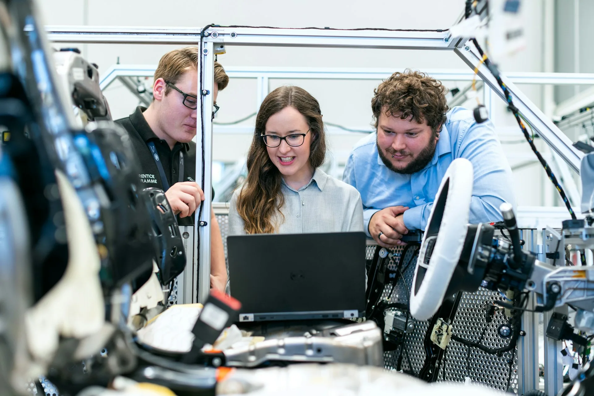 Three engineers share one laptop at a vehicle test rig with a steering wheel and wiring harnesses around them.