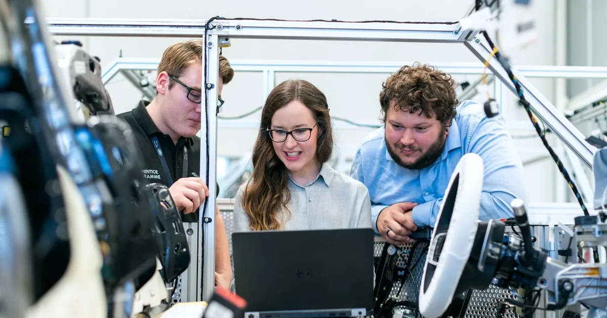 Three engineers look at a laptop together beside a vehicle test rig with a steering wheel and wiring in a development workshop.